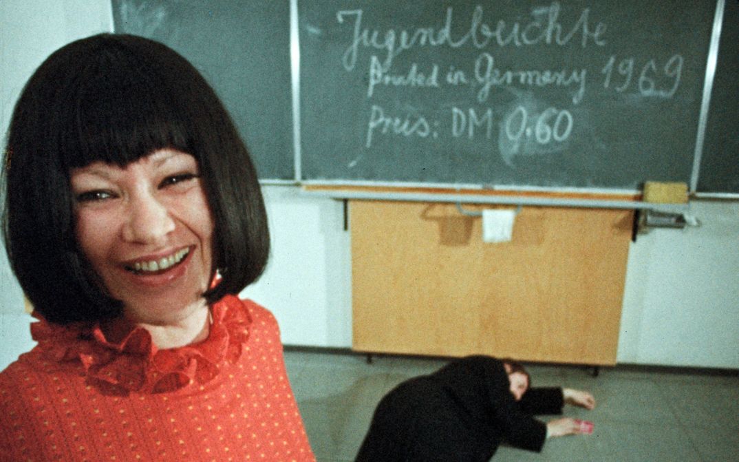 Filmstill aus GESCHICHTEN VOM KÜBELKIND: Eine Frau mit schwarzem Haar und rotem Oberteil lacht in die Kamera. Hinter ihr ist eine Wandtafel zu sehen, auf dem Boden liegt ein schwarz angezogener Mann.