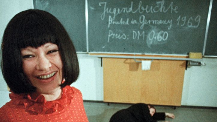 Filmstill aus GESCHICHTEN VOM KÜBELKIND: Eine Frau mit schwarzem Haar und rotem Oberteil lacht in die Kamera. Hinter ihr ist eine Wandtafel zu sehen, auf dem Boden liegt ein schwarz angezogener Mann.