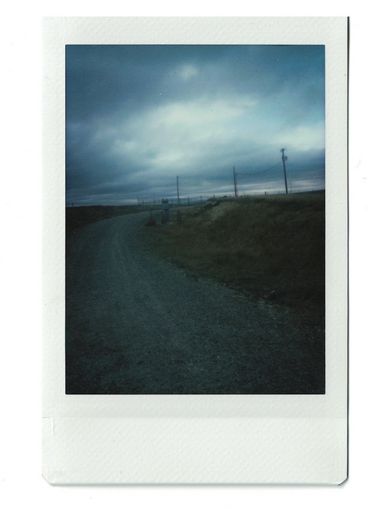 Polaroid of a dark gravel road, with a stormy sky in the background.