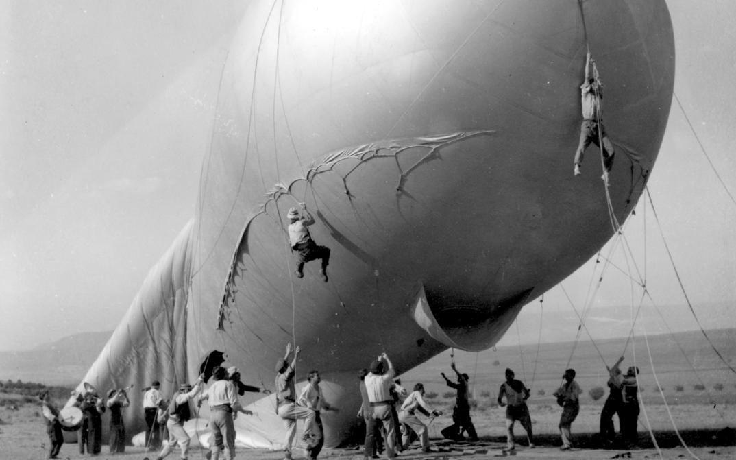 Men hold the ropes of a large barrage balloon, two of them hovering in the air.Film still from THE ATTACHED BALLOON. 
