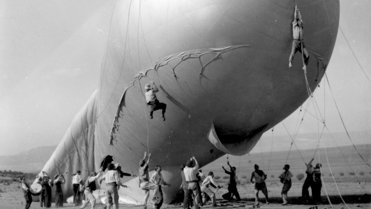 Men hold the ropes of a large barrage balloon, two of them hovering in the air.Film still from THE ATTACHED BALLOON. 