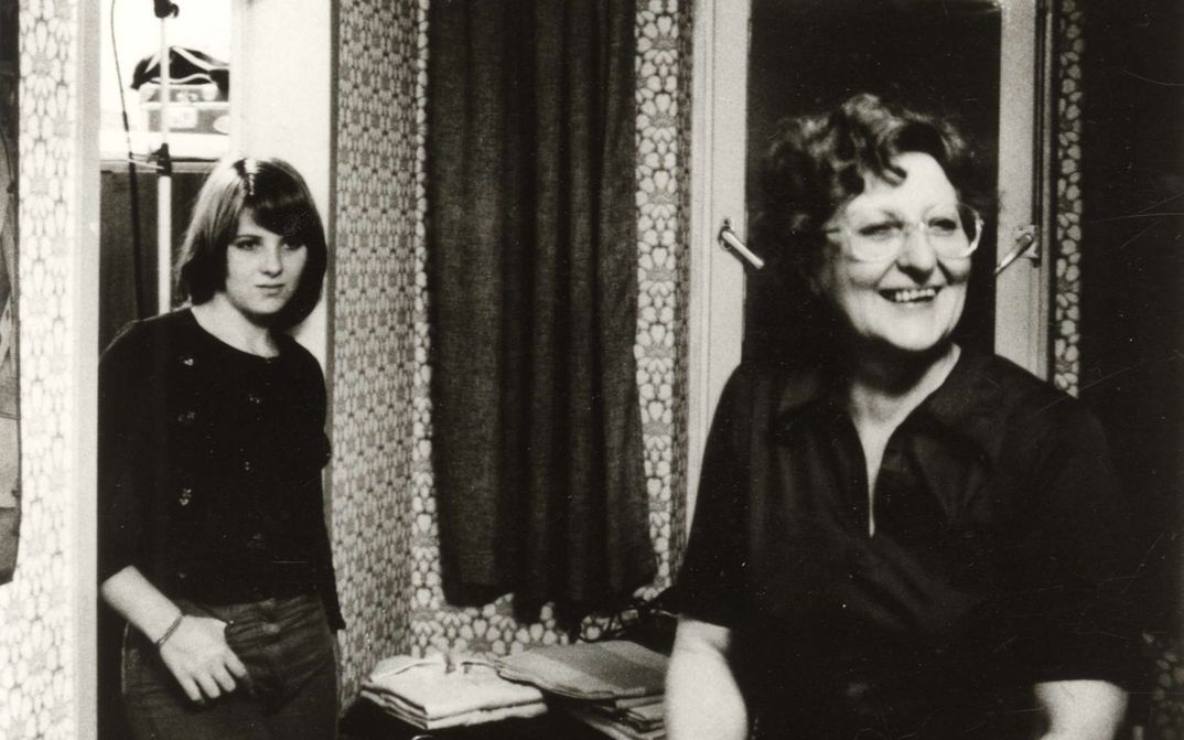 Filmstill aus VON WEGEN SCHICKSAL: Eine Frau steht lachend im Wohnzimmer, an den Türrahmen lehnt sich eine Teenagerin.