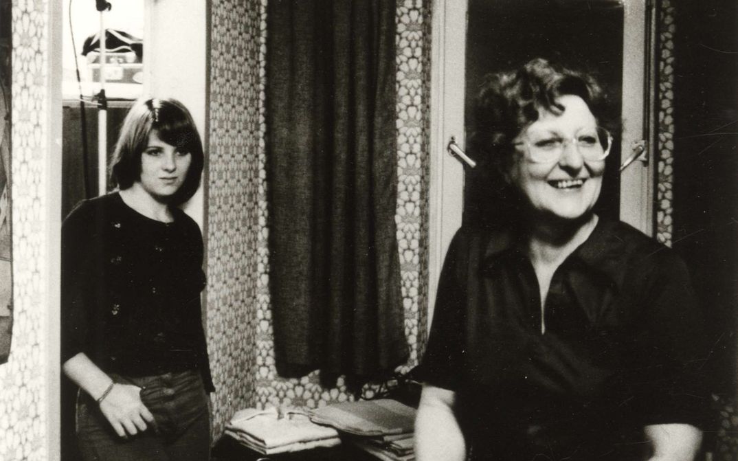 Filmstill aus VON WEGEN SCHICKSAL: Eine Frau steht lachend im Wohnzimmer, an den Türrahmen lehnt sich eine Teenagerin.