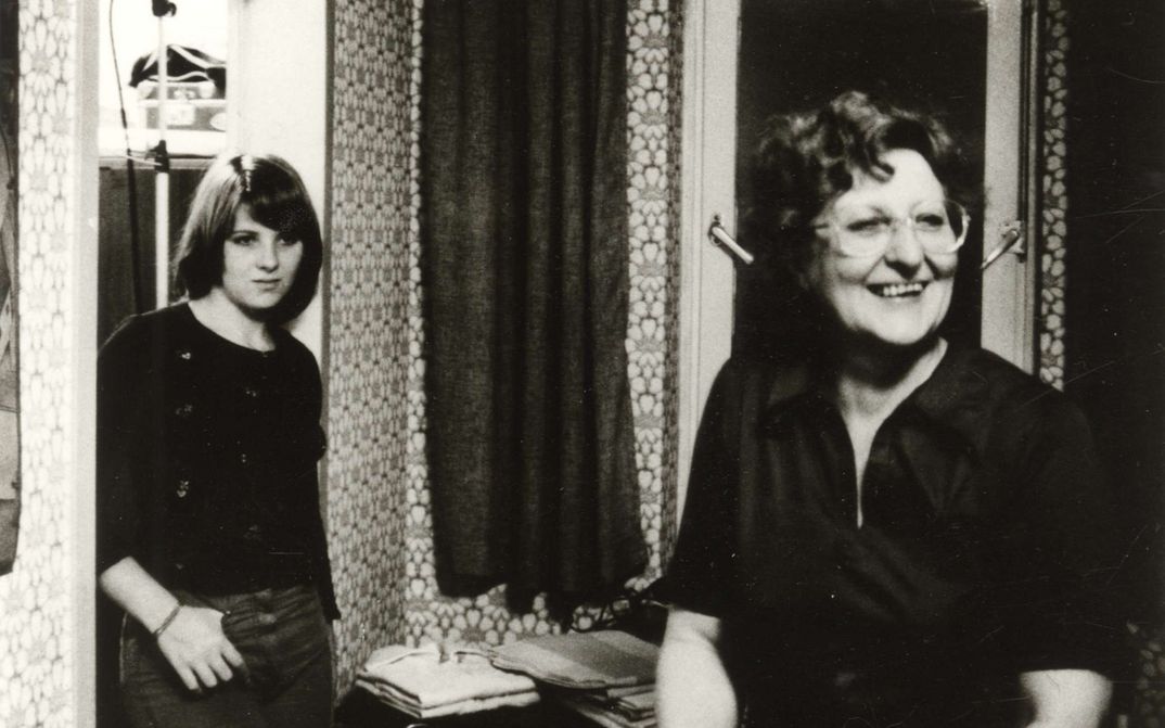 Filmstill aus VON WEGEN SCHICKSAL: Eine Frau steht lachend im Wohnzimmer, an den Türrahmen lehnt sich eine Teenagerin.