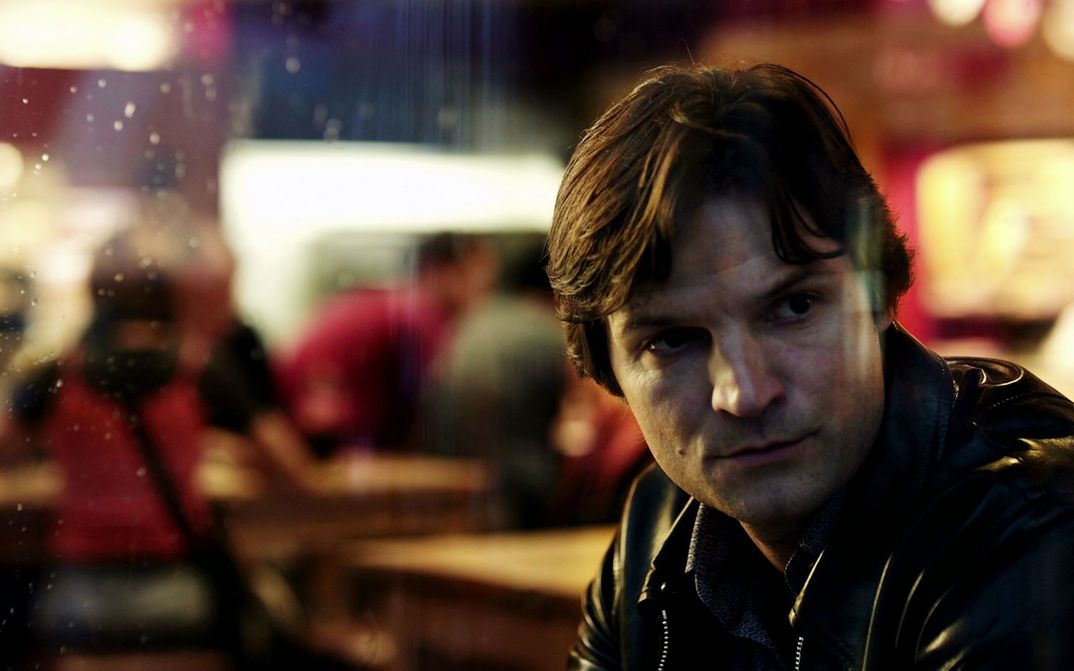 Film still from IM SCHATTEN: A man is sitting alone in a bar. You can see him through the glass, he is looking out.