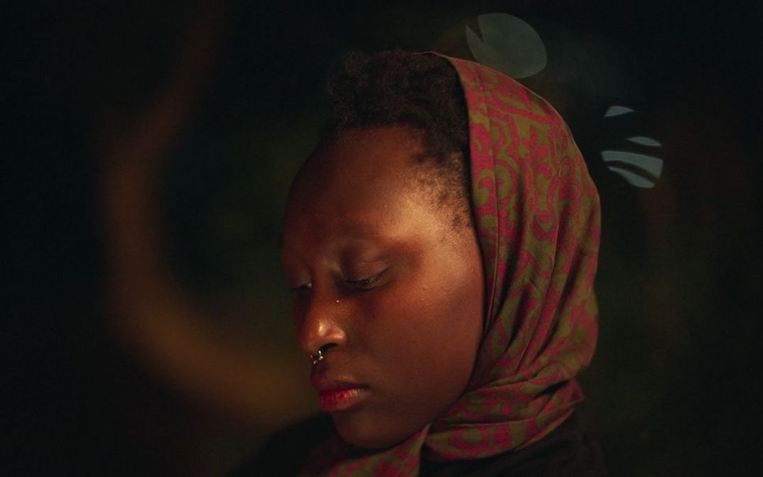 A close-up of a young woman wearing a green and red patterned headscarf against a dark background. 