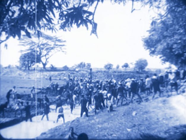 Film still from Chanasorn Chaikitiporn’s film “Here We Are”. A cyanotype-style image of a large group of people walking through a nature path. 