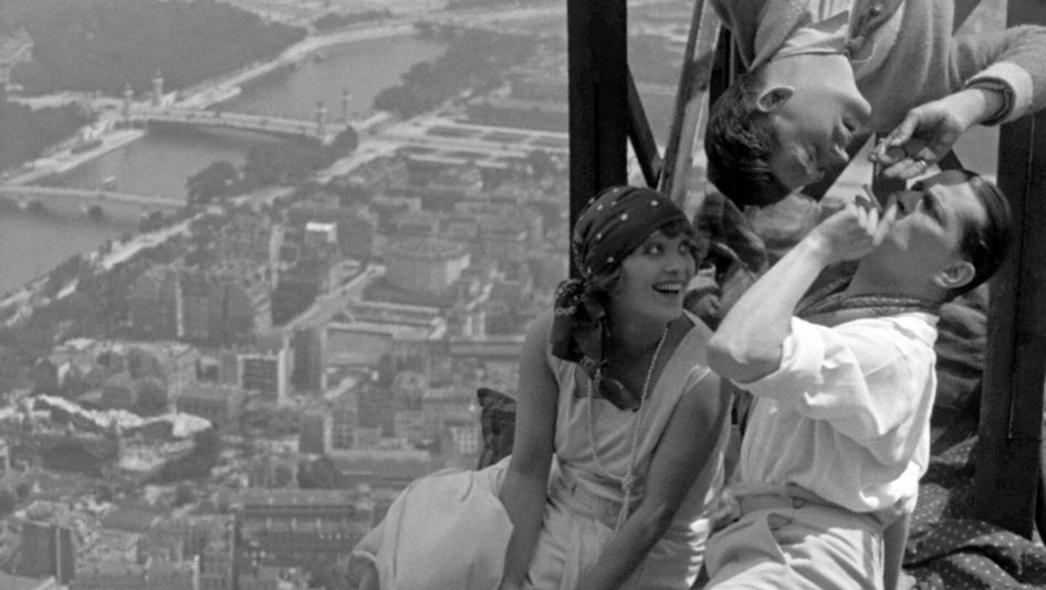 Film still from PARIS QUI DORT: Three young, boisterous people are sitting in the Eiffel Tower. Below them, the city looks tiny.