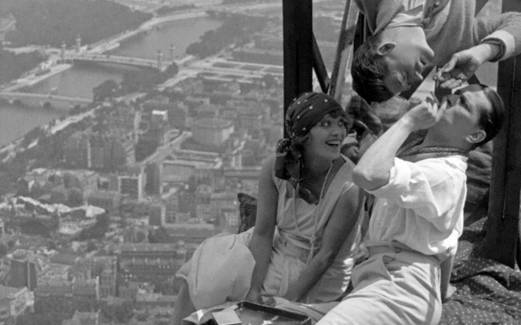 Filmstill aus PARIS QUI DORT: Drei junge, ausgelassene Menschen sitzen auf dem Eiffelturm. Unter ihnen sieht man klein die Stadt.