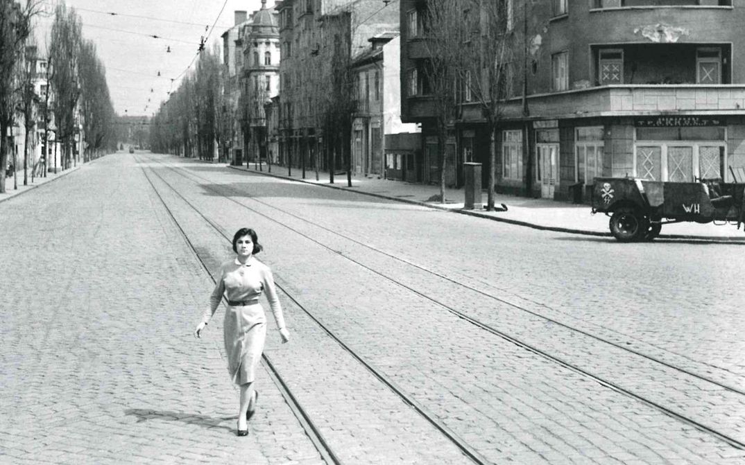 Filmstill aus WE WERE YOUNG: Eine menschenleere Straße mit Tramgleisen. Eine junge Frau geht mit entschlossenem Ausdruck quer über die Straße.