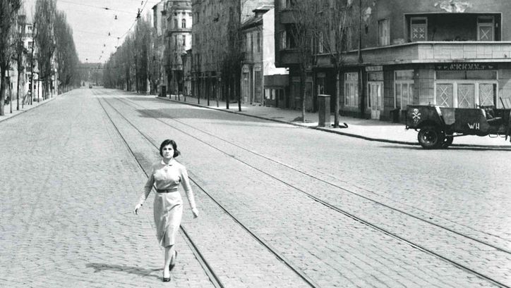 Filmstill aus WE WERE YOUNG: Eine menschenleere Straße mit Tramgleisen. Eine junge Frau geht mit entschlossenem Ausdruck quer über die Straße.