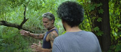 Still from the film "Camuflaje (Camouflage)" by Jonathan Perel. Two men in an overgrown forest. One holds out his arms, as if to show how big something is. The other watches, we only see his back.