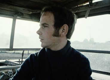 Still from the film "Ein Herbst im Ländchen Bärwalde" by Gautam Bora. A man in a blue turtleneck sweater is looking to the left. He seems to be sitting in a car, there are windows behind him and a hatch is slightly open above him.