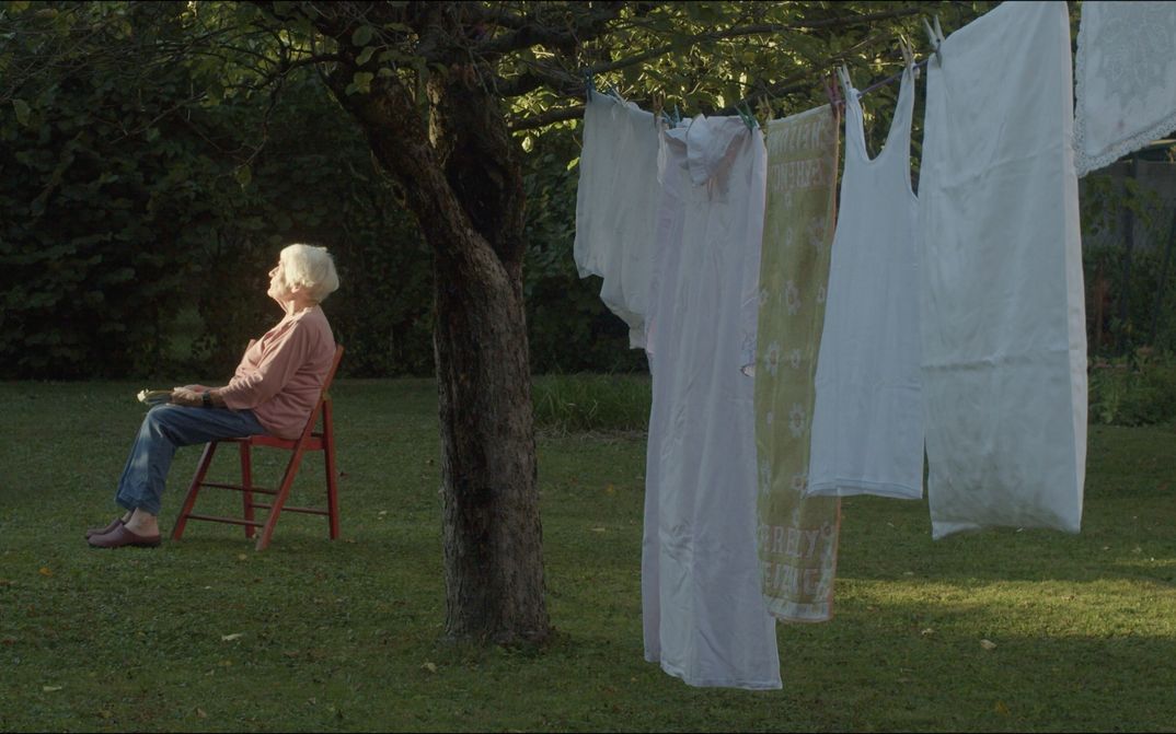 Film still from DER TAG VOR DEM ABEND: An elderly woman with white hair is sitting on a chair in a garden, gazing at the sun. In the foreground, laundry hangs on a line tied to a tree.