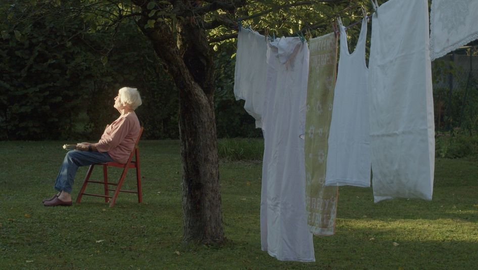 Filmstill aus DER TAG VOR DEM ABEND: Eine alte Frau mit weißen Haaren sitzt in einem Garten auf einem Stuhl und blickt in die Sonne. Im Vordergrund hängt Wäsche an einer Leine, die an einem Baum befestigt ist.