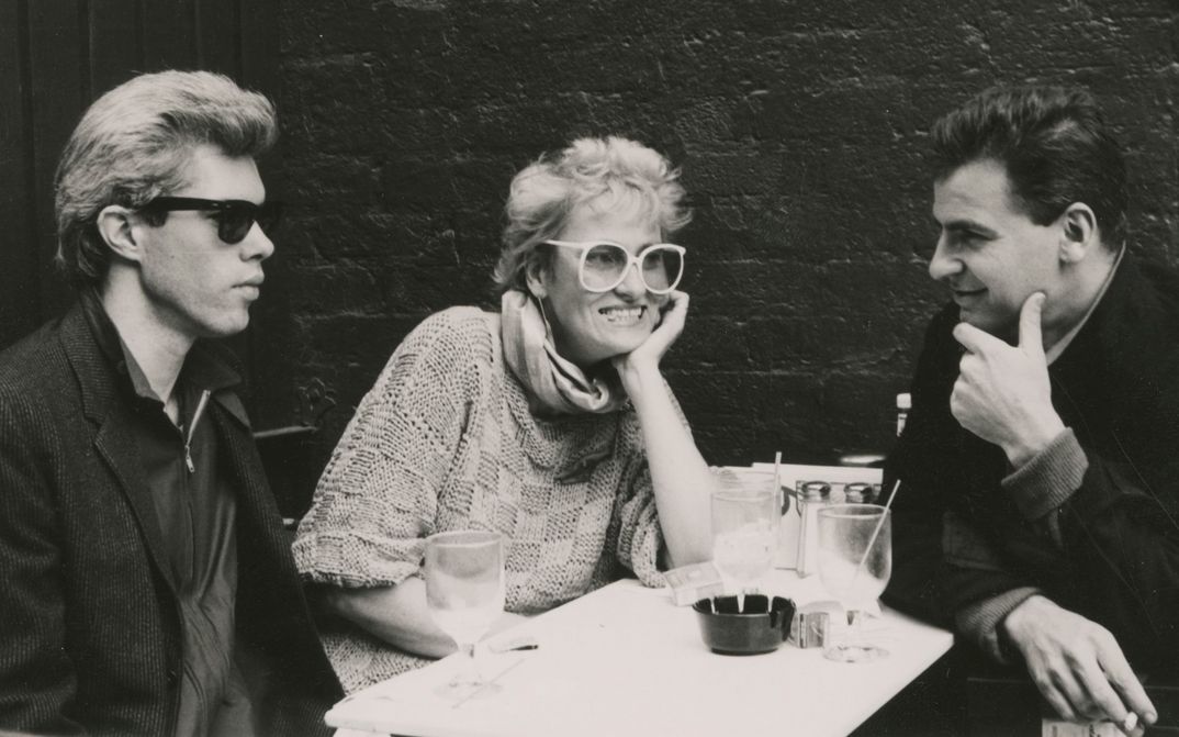 Filmstill from FRÄULEIN BERLIN: Two men and a woman are sitting at a table. They have drinks, wear large glasses and are partly smoking.