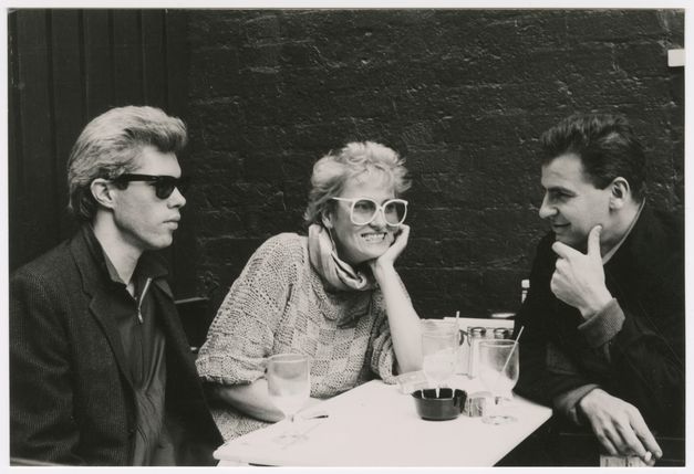 Filmstill aus FRÄULEIN BERLIN: Zwei Männer und eine Faru sitzen an einem Tisch. Sie haben Getränke, tragen große Brillen und rauchen teils.