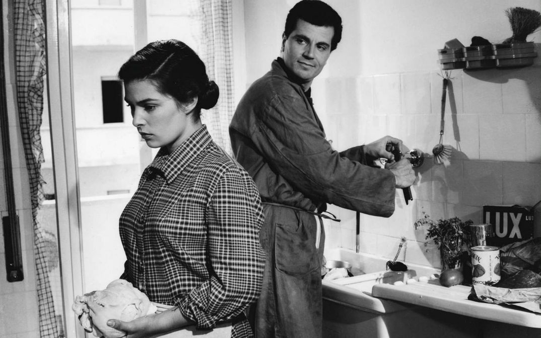 Film still from IL SOLE NEGLI OCCHI: A young woman and a man are standing in a kitchen, both working and turning their backs on each other. He is half turned towards her and looking at her.