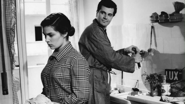 Film still from IL SOLE NEGLI OCCHI: A young woman and a man are standing in a kitchen, both working and turning their backs on each other. He is half turned towards her and looking at her.