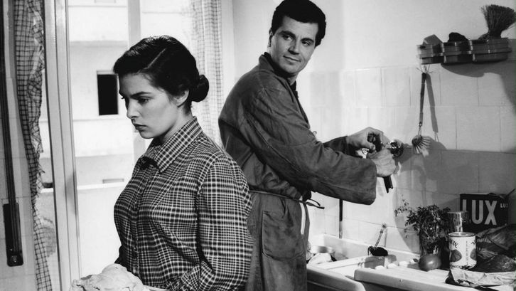 Filmstill aus IL SOLE NEGLI OCCHI: Eine junge Frau und ein Mann stehen in einer Küche, beide arbeitend und einander den Rücken zukehrend. Er ist halb zu ihr umgedreht und schaut sie an.