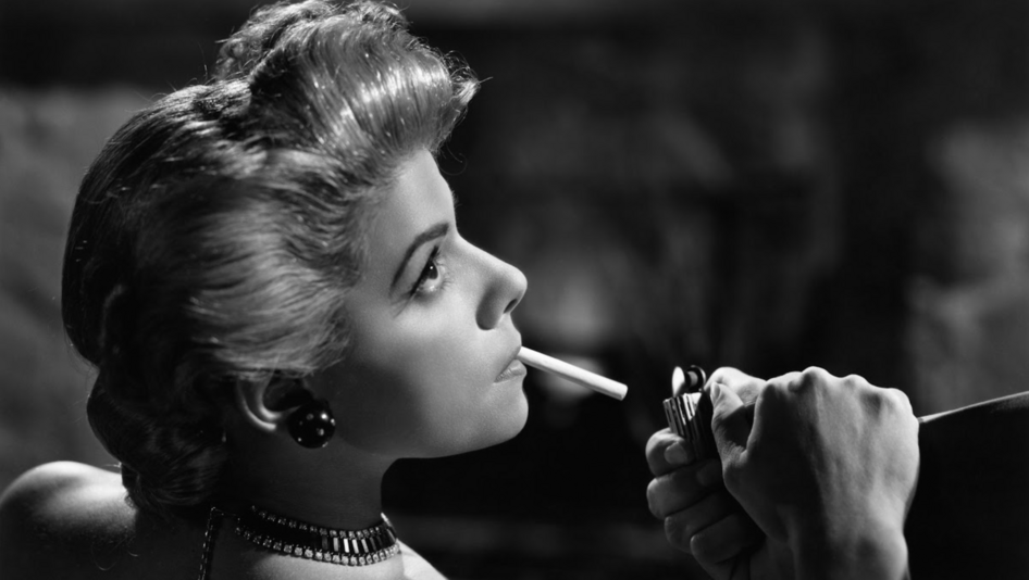 Film still from LLÉVAME EN TUS BRAZOS: An elegantly dressed and coiffed woman has someone light her cigarette.