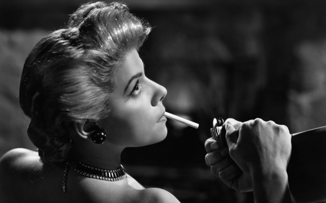 Film still from LLÉVAME EN TUS BRAZOS: An elegantly dressed and coiffed woman has someone light her cigarette.