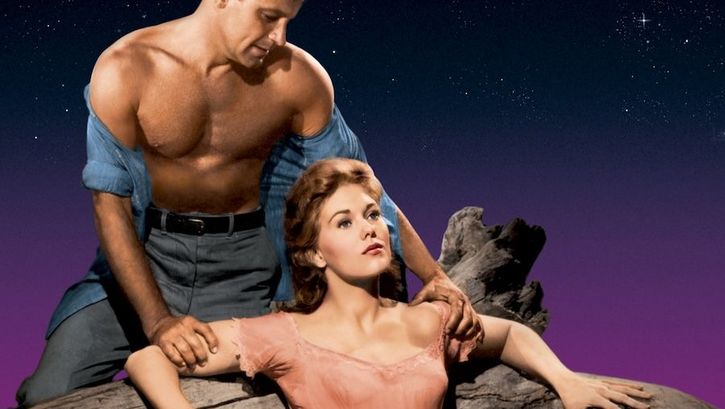 Filmstill aus PICNIC: Ein muskulöser Mann, dessen offenes Hemd von den Schultern gerutscht ist, greift die Arme einer vor ihm sitzenden Frau.