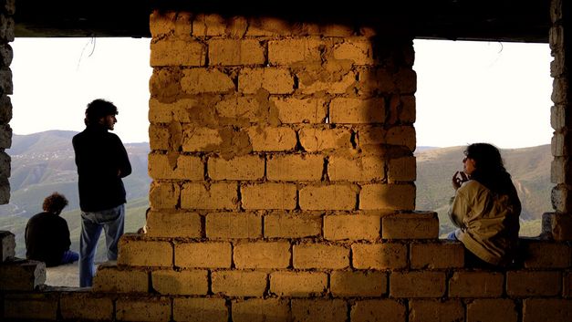 Film still from INNER BLOOMING SPRINGS by Tiku Kobiashvili. The image shows an unplastered wall with openings through which three people and a hilly landscape can be seen.  Tiku Kobiashvili. Das Bild zeigt eine unverputzte Mauer mit Durchbrüchen, durch die hindurch man drei Personen und eine hügelige Landschaft sieht.