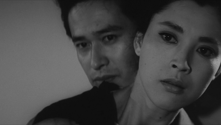 Film still from WOMAN OF THE LAKE: We see the heads of a woman and a man. He is standing behind her, both looking towards the camera.