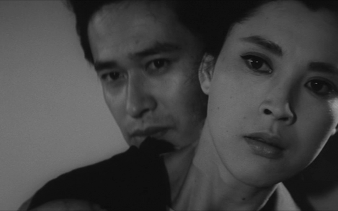 Film still from WOMAN OF THE LAKE: We see the heads of a woman and a man. He is standing behind her, both looking towards the camera.