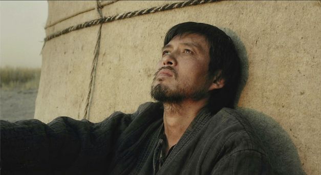 Still from the film "Akyn (Poet)" by Darezhan Omirbayev. A bearded man sits against a structure, looking upward.