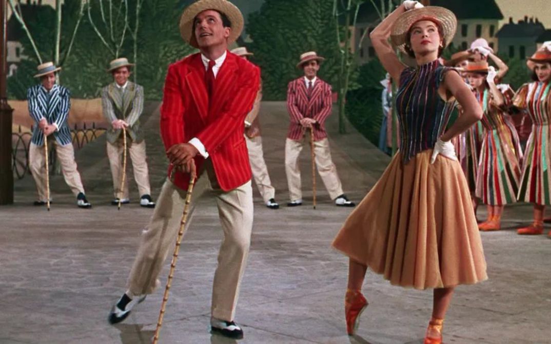 Filmstill aus AN AMERICAN IN PARIS: Eine Tanzszene in einer Studio-Kulisse: Im Vordergrund tanzen ein Mann und eine Frau in sommerlich-bunter Kleidung. Im Hintergrund weitere Tänzerinnen und Tänzer.