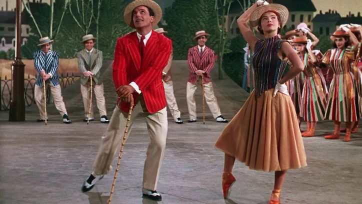 Filmstill aus AN AMERICAN IN PARIS: Eine Tanzszene in einer Studio-Kulisse: Im Vordergrund tanzen ein Mann und eine Frau in sommerlich-bunter Kleidung. Im Hintergrund weitere Tänzerinnen und Tänzer.