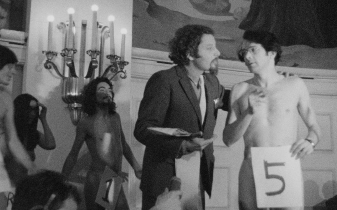 Film Still aus dem Film „Exprmntl 4 Knokke“ von Claudia von Alemann. Schwarzweißes Bild einer Performance in einem Innenraum: Mehrere nackte Männer stehen nebeneinander, nummerierte Schilder („1“ im Hintergrund, „5“ im Vordergrund) bedecken jeweils ihre Genitalien, während ein Mann im Anzug wie ein Moderator mit einem der Teilnehmer spricht; im Vordergrund dokumentiert ein Reporter mit Aufnahmegerät die Szene, im Hintergrund sind Kerzenleuchter und Wandbilder zu sehen.