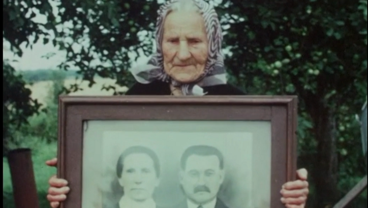Film still from GOING HOME. An old woman holds up a framed portrait photograph of a couple. 
