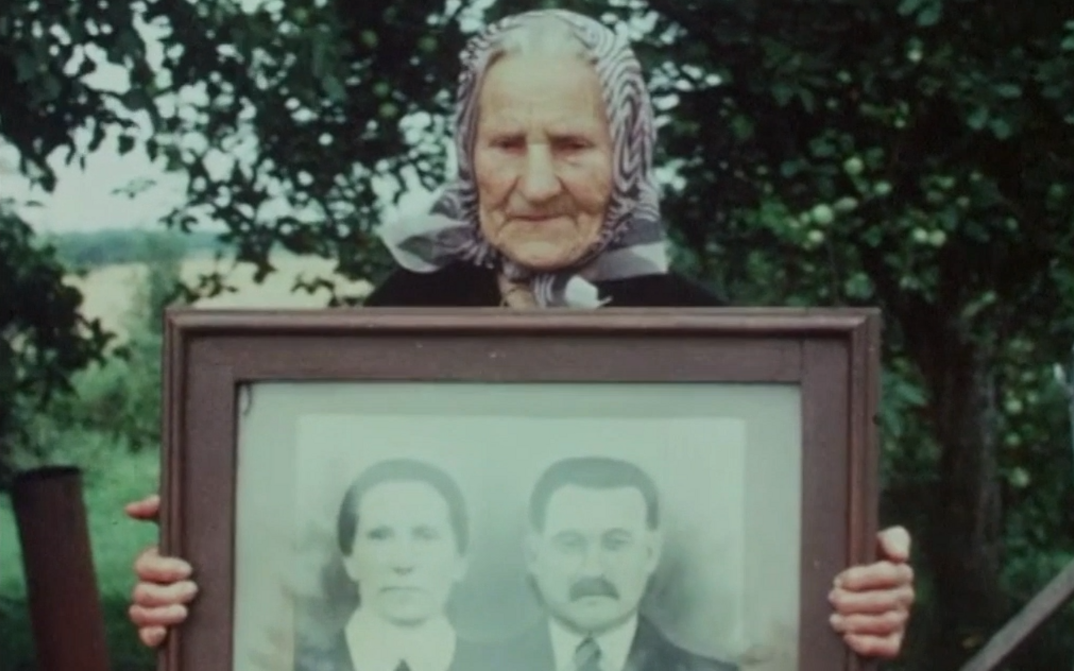 Filmstill aus GOING HOME. Eine alte Frau hält eine gerahmte Porträtfotografie eines Paares hoch. 