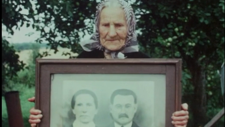Film still from GOING HOME. An old woman holds up a framed portrait photograph of a couple. 