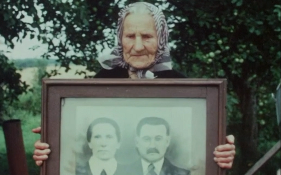 Film still from GOING HOME. An old woman holds up a framed portrait photograph of a couple.