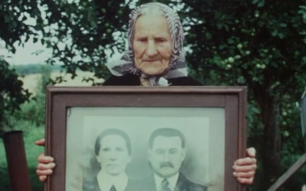 Film still from GOING HOME. An old woman holds up a framed portrait photograph of a couple.