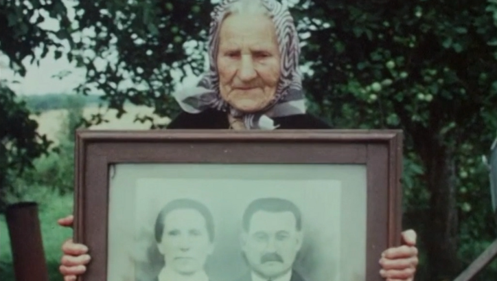 Filmstill aus GOING HOME. Eine alte Frau hält eine gerahmte Porträtfotografie eines Paares hoch. 