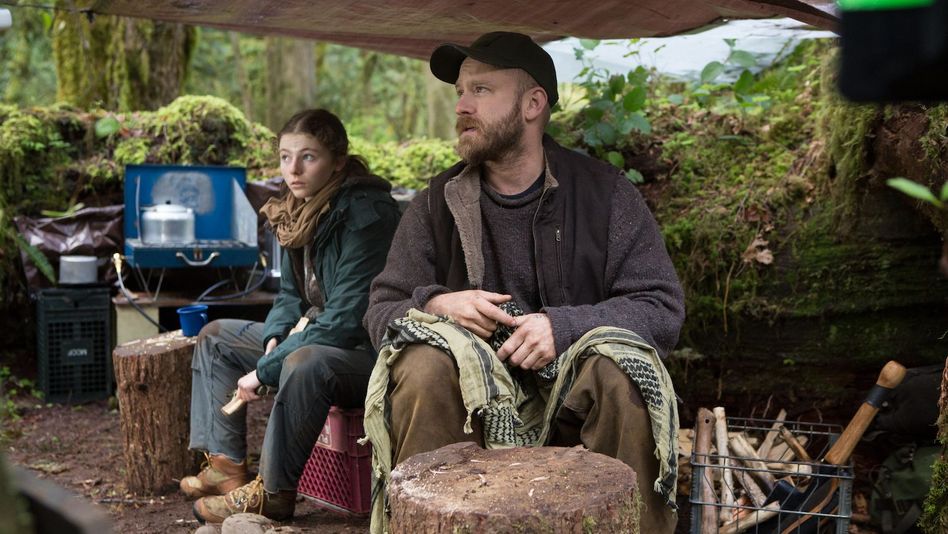 Film still from LEAVE NO TRACE: A man and a teenage girl are sitting under a tarp in the woods. Cooking gear can be seen in the background.