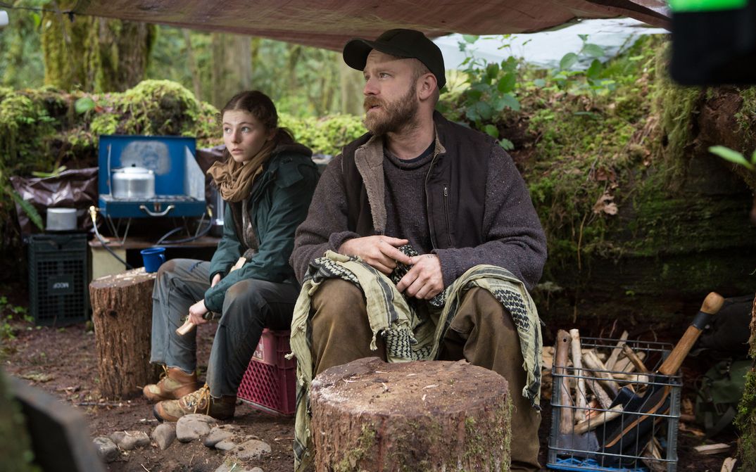 Film still from LEAVE NO TRACE: A man and a teenage girl are sitting under a tarp in the woods. Cooking gear can be seen in the background.
