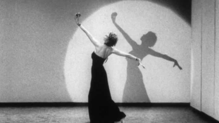 Filmstill aus LIVES OF PERFORMERS: Eine tanzende Frau in einem langen Kleid in einem Lichtkegel.