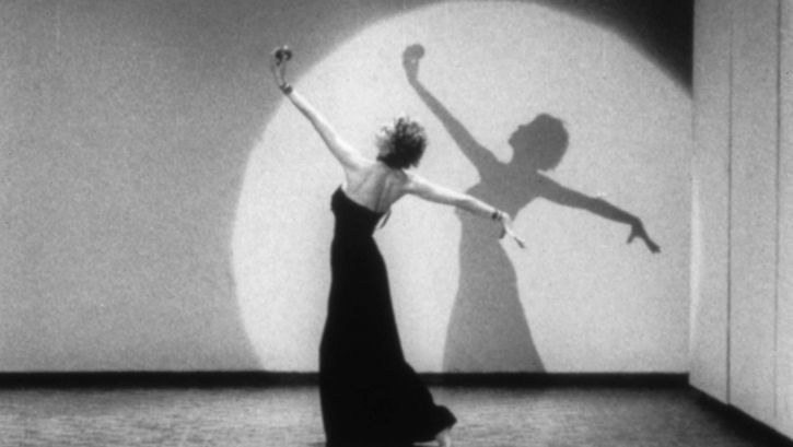 Film still from LIVES OF PERFORMERS: A dancing woman in a long dress in a cone of light.