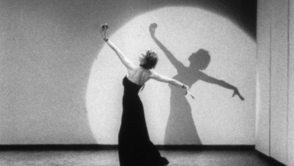 Film still from LIVES OF PERFORMERS: A dancing woman in a long dress in a cone of light.