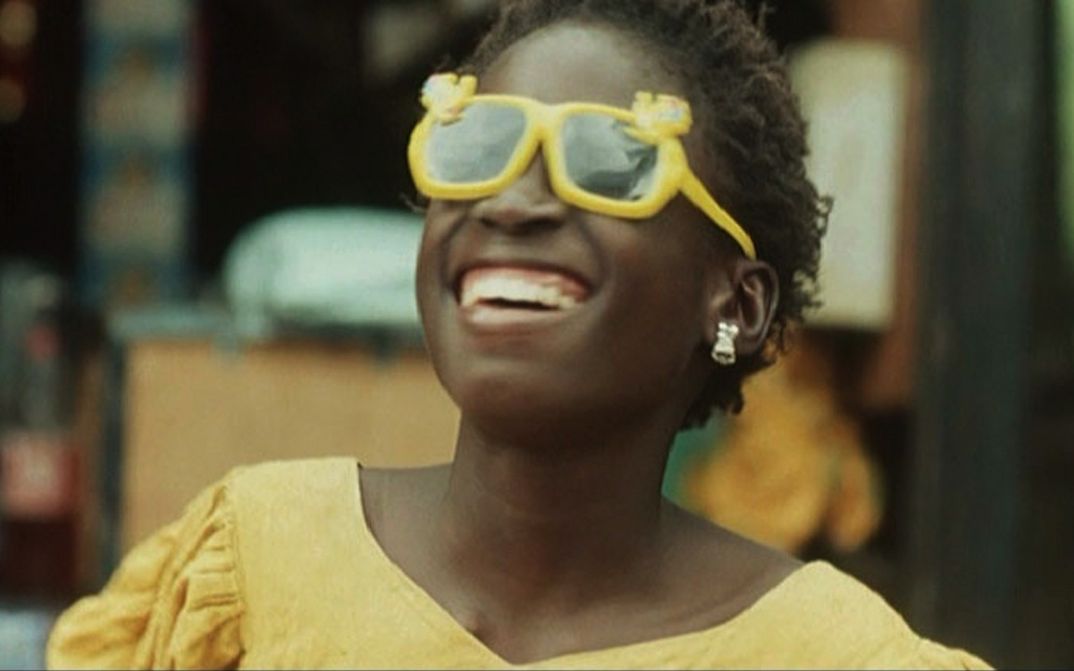 Filmstill aus LA PETITE VENDEUSE DE SOLEIL: Ein Mädchen in einem gelben Kleid und einer gelben Sonnenbrille schaut Richtung Himmel und lacht dabei.