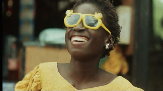 Film still from LA PETITE VENDEUSE DE SOLEIL: A girl in a yellow dress and yellow sunglasses looks up at the sky and laughs.