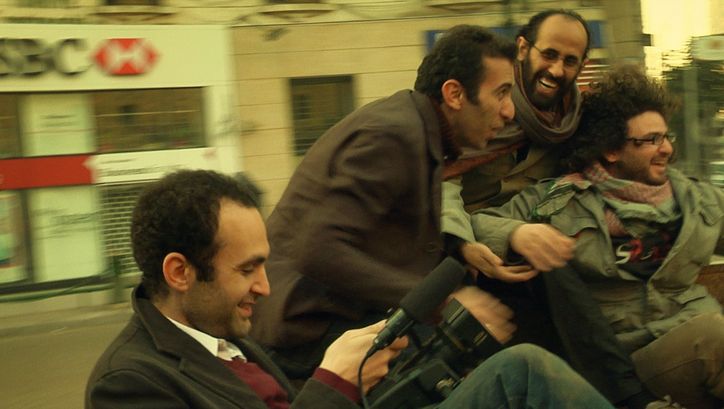 Film still from IN THE LAST DAYS OF THE CITY: Four men laughing with a camera on the street. The background is blurred.