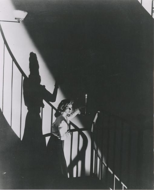 Film still from THE SPIRAL STAIRCASE: A young woman walks down a spiral staircase with a candle in her hand. The light casts dramatic shadows.
