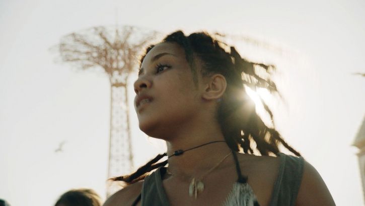 Filmstill aus TENDABERRY: Man sieht eine junge Frau, aus der Untersicht und leicht unscharf aufgenommen, an einem sonnigen Tag.